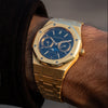 A gold-plated Audemars Piguet Royal Oak wristwatch with a blue dial and moonphase subdials, shown worn on a person's wrist with a black sleeve visible.